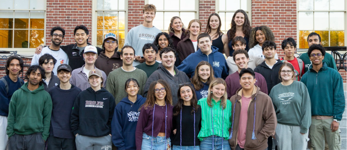 A large group of young adults posing together on outdoor steps in front of a brick building with large windows.